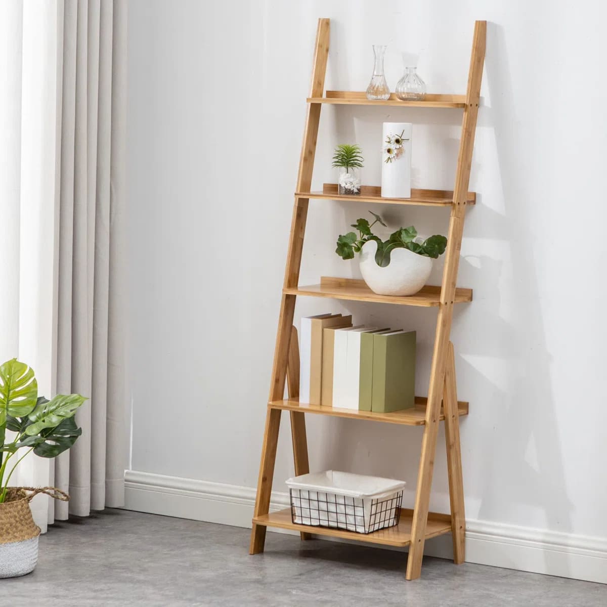 A wooden ladder shelf stands against a white wall, offering stylish storage for renters. It holds books, potted plants, decorative vases, and a wire basket. A green leafy plant in a basket sits on the floor nearby, with light curtains hanging to the left.