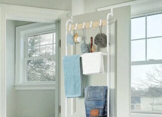 An over-the-door organizer offers smart storage for renters, hanging on a white bathroom door and holding towels, clothes, and toiletries. A toilet, window, and bathtub are visible in the background with dark tile flooring.