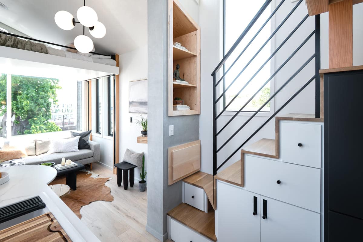Modern, bright tiny house interior with a loft bed, cozy living area, built-in wooden shelving and storage under the staircase, large windows, and minimalist decor. Natural light fills the space, highlighting its clever design.