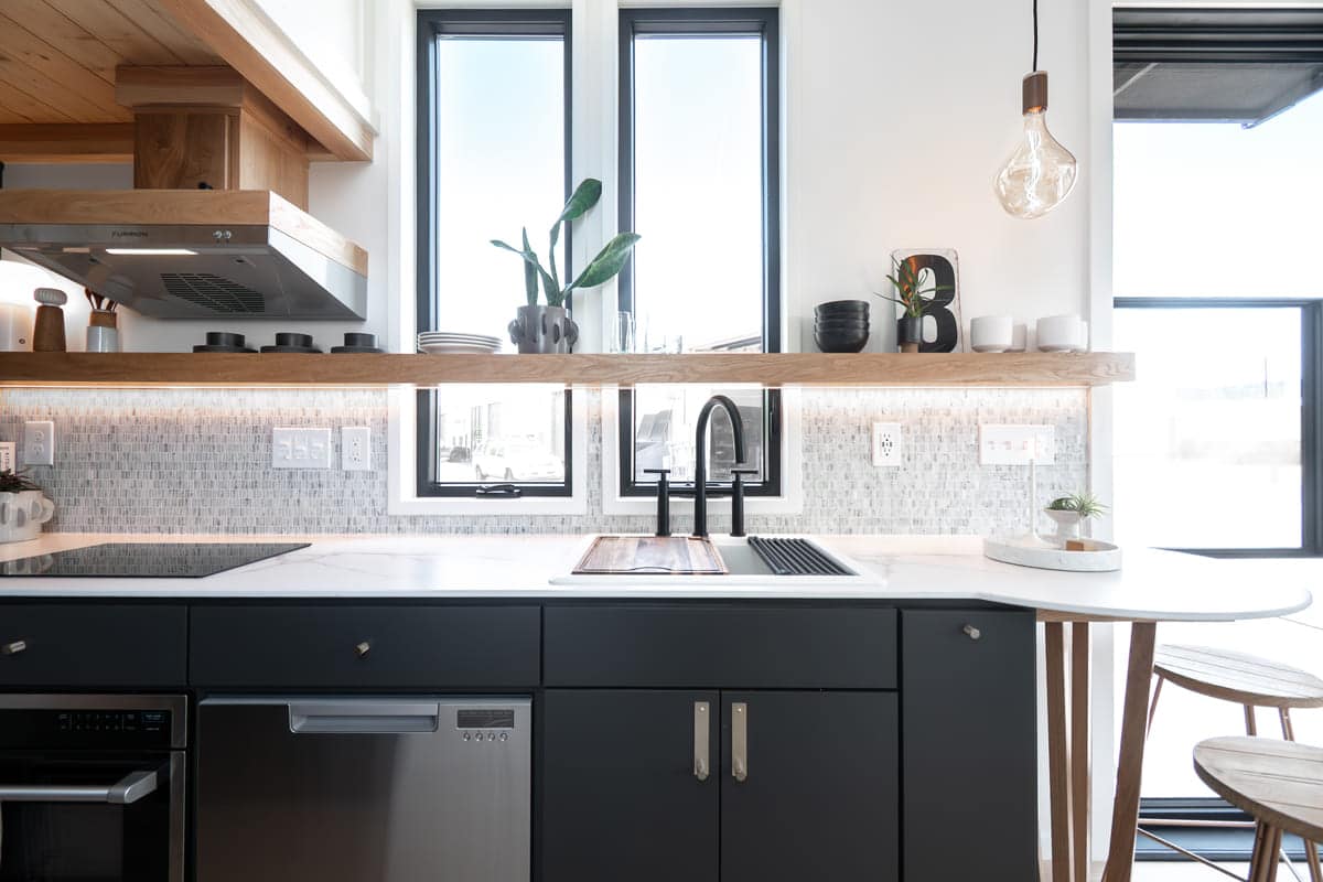Modern tiny house kitchen with black lower cabinets, white countertops, a sleek black faucet, mosaic tile backsplash, wooden shelves with plants and dishes, two large windows, and pendant lighting.