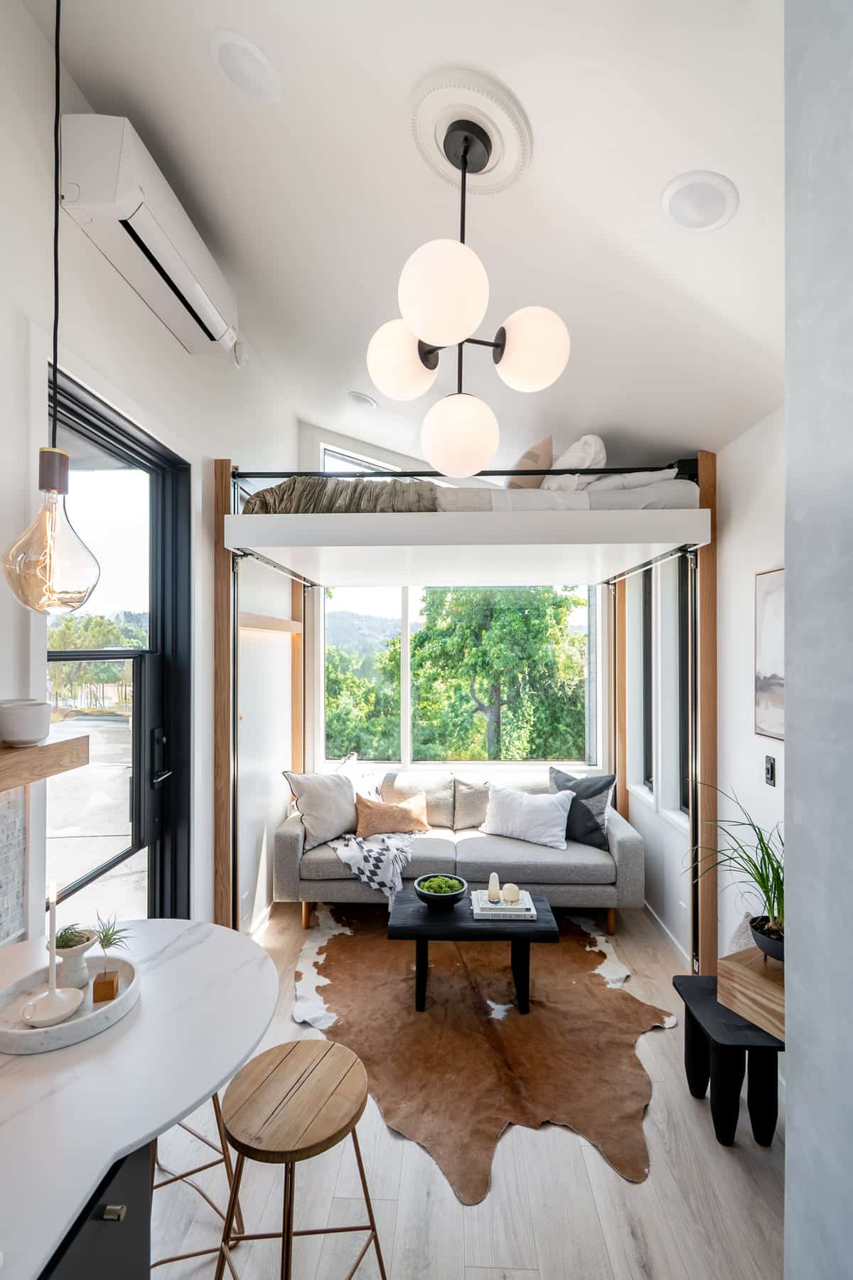 A bright, modern tiny house living room with a gray sofa, cowhide rug, coffee table, and large windows. Above is an elevator bed with cozy bedding. The space features stylish lighting, wood accents, and green trees visible outside.