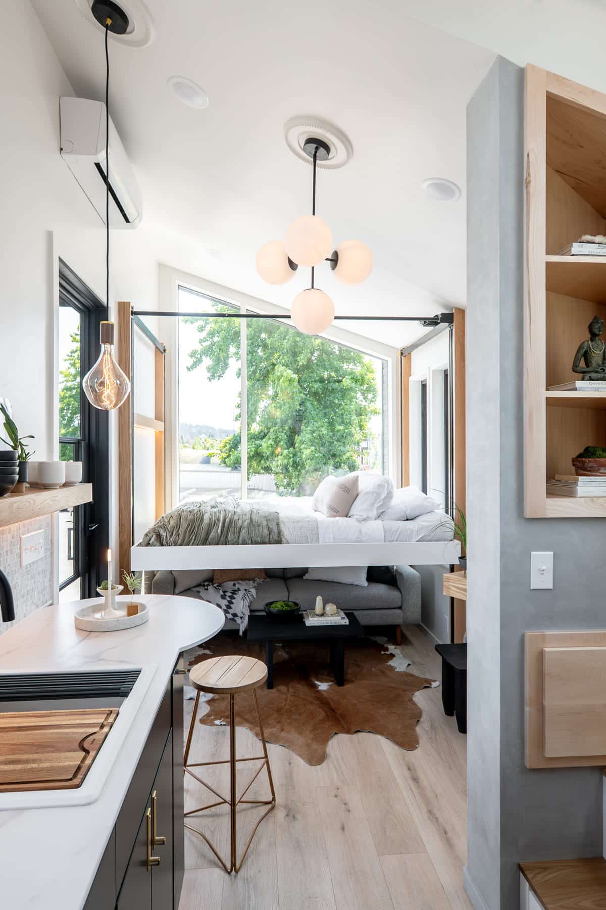 A modern tiny house interior with an elevator bed beside large windows, a small kitchen with a bar stool, light wood floors, a cowhide rug, and minimalistic decor. Natural light fills the bright, airy space.