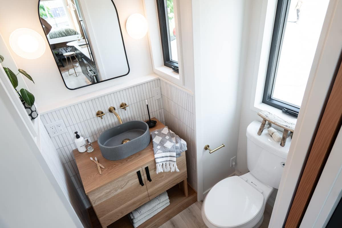 Modern small bathroom in a tiny house with a round mirror, wall-mounted lights, gray vessel sink on a wooden vanity, towel, and white toilet next to a window with decor. Light wood and white tile accents throughout.