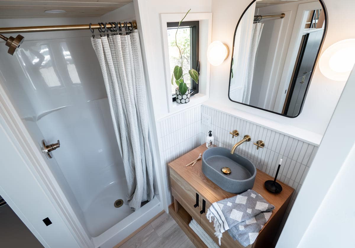 Modern tiny house bathroom with a corner shower, white curtain, wooden vanity with a gray basin sink, gold faucet, round mirror, wall lights, and a small window with a potted plant. Gray towel folded on the counter.