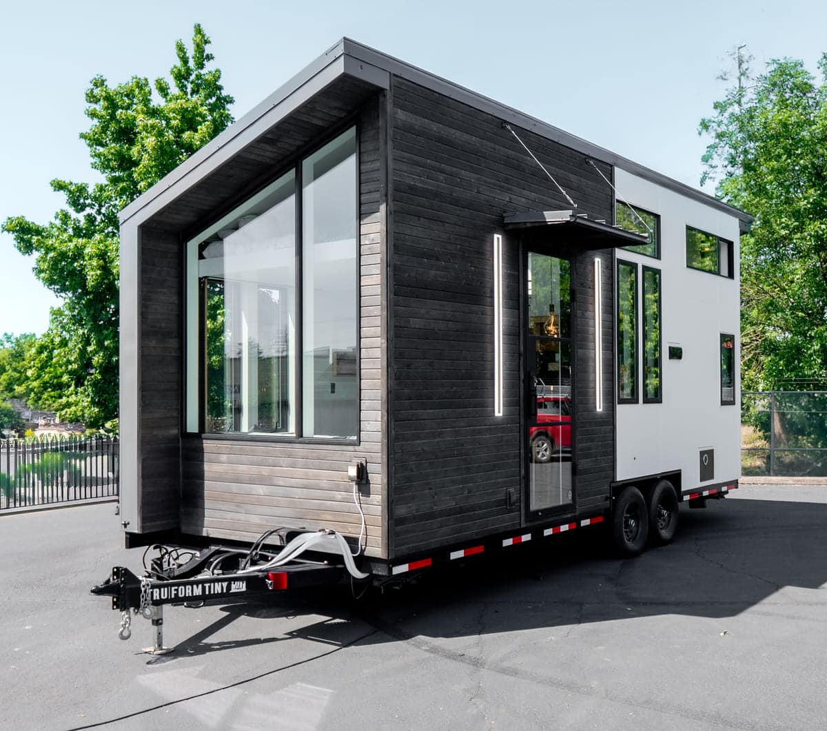 Modern tiny house on wheels with large windows and dark wood siding, parked on pavement near trees and a fence. The sleek, minimalist design includes a visible side door and features an innovative elevator bed inside.