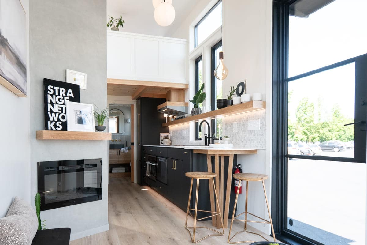 Modern tiny house interior with light wood floors, black kitchen cabinets, a long countertop with stools, wall-mounted shelves, plants, and large windows letting in natural light. An elevator bed, fireplace, and artwork decorate the small living space.