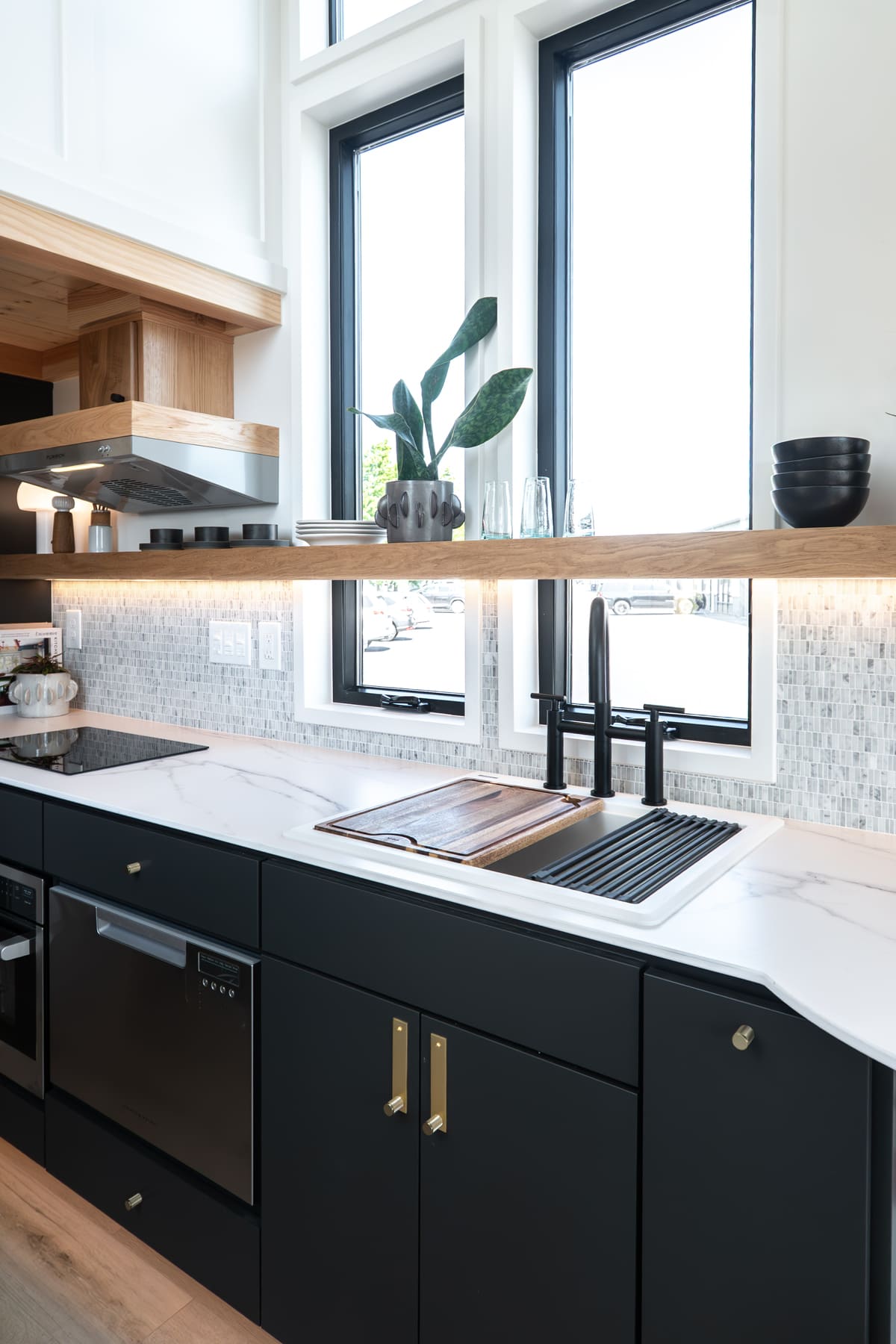 Modern tiny house kitchen with black cabinets, brass handles, white marble countertop, a black sink with faucet, wooden shelf for dishes and plants, and tall windows filling the space with natural light—perfect beneath an elevator bed.