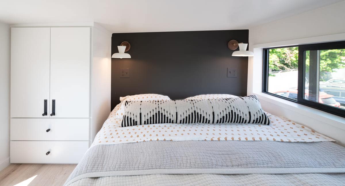 A neatly made elevator bed with patterned pillows and a gray quilt is set against a black headboard with two white wall-mounted reading lamps, making this tiny house bedroom feel bright and cozy, thanks to a large window letting in natural light.