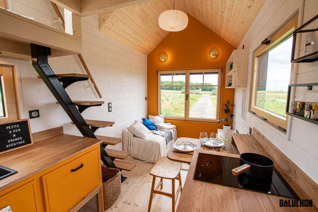 A cozy, modern tiny house interior features white and orange walls, wood accents, a small kitchen, a loft bed, a couch with pillows, large windows, and a ladder-style staircase. Natural light fills this charming tiny house space.