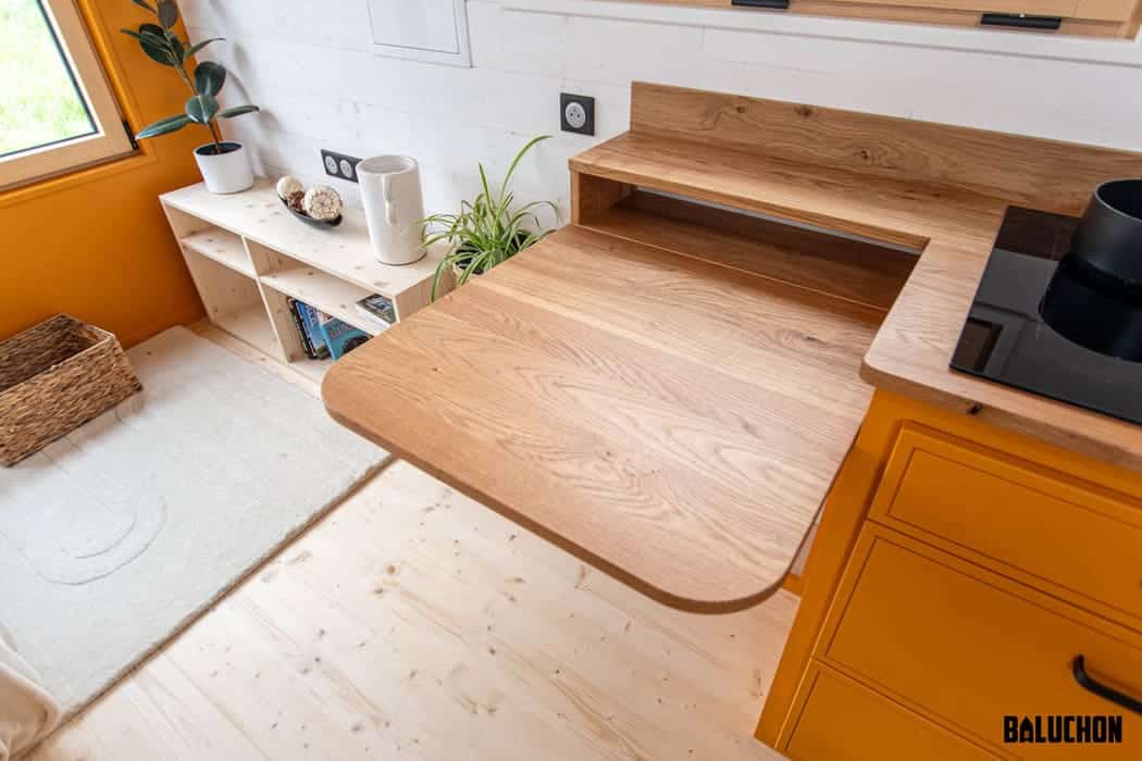 A small kitchen in a tiny house with bright orange cabinets features a wooden pull-out countertop extension, next to a black stovetop and plants on light wood flooring. A wicker basket and shelves are visible in the background.