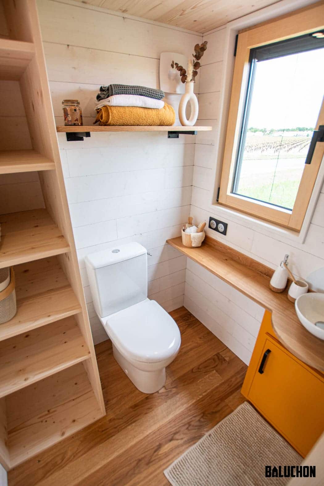 Small, modern bathroom in a tiny house with wood accents, a white toilet, open shelving, a counter with a vessel sink, a window, and a shelf holding towels and decorative items. Natural light fills the space.