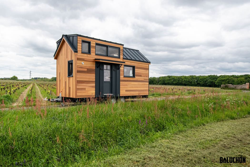 A stylish tiny house with wood panel siding and black trim sits on a grassy field beside rows of crops under a cloudy sky. The BALUCHON logo is visible in the bottom right corner.