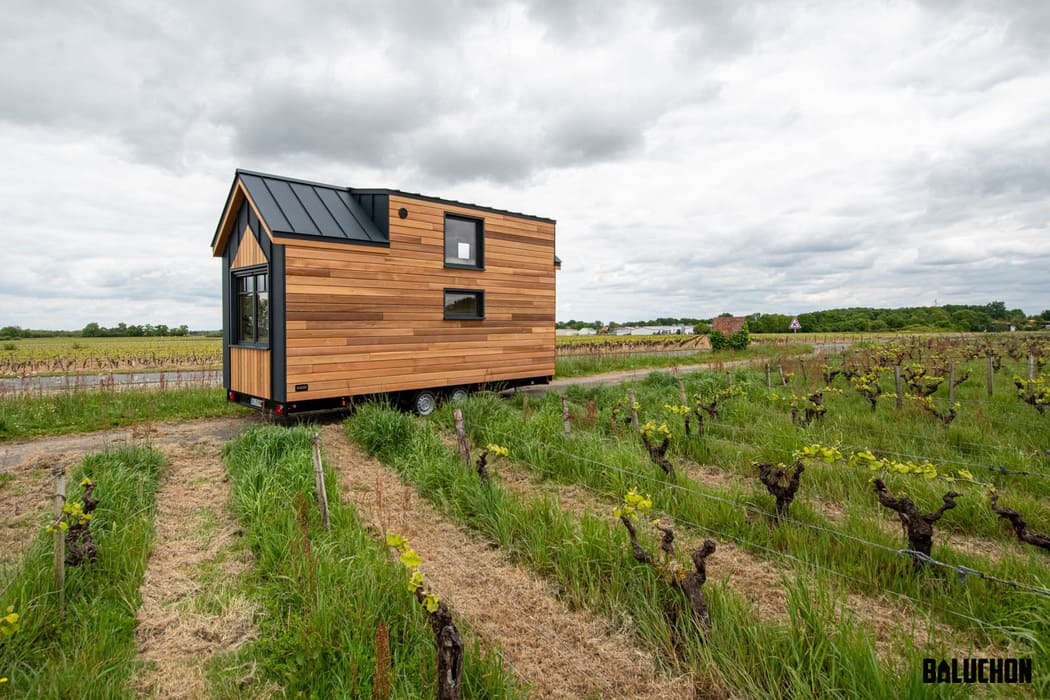 A modern tiny house on wheels sits in the middle of a green vineyard under a cloudy sky. With large windows and a sleek black metal roof, this wooden retreat showcases the Baluchon logo in the lower right corner.