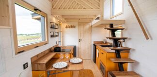 A modern tiny house kitchen with orange cabinets, wooden countertops, a cozy dining area set for two, open shelves, and a unique wooden staircase leading to the loft. A potted plant brightens up the corner.