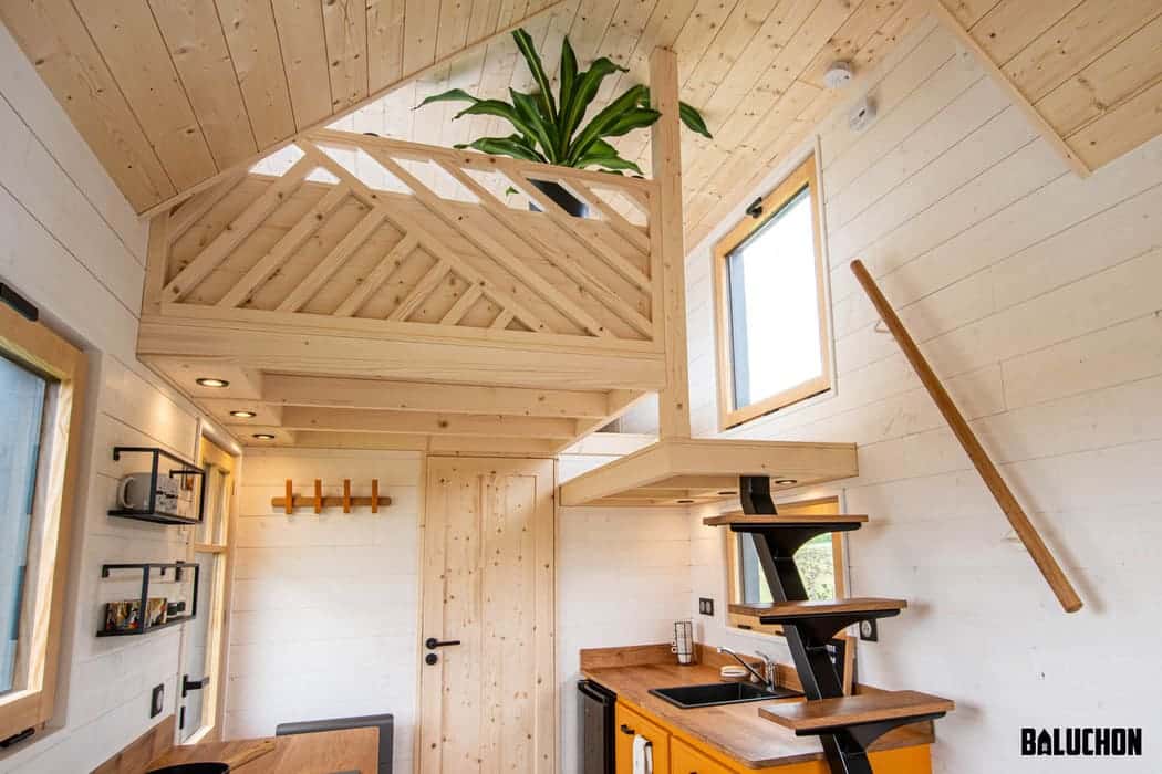 A modern tiny house interior featuring light wood walls and ceiling, black accents, a compact kitchen with open shelves, a loft adorned with a plant, and a minimalist wooden staircase leading to the cozy loft.