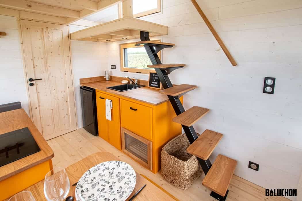 A bright, modern tiny house kitchen with yellow cabinets, a small black fridge, wood countertops, and a minimalist wooden staircase leading to a loft. Light wood walls and flooring create a cozy, open tiny house atmosphere.