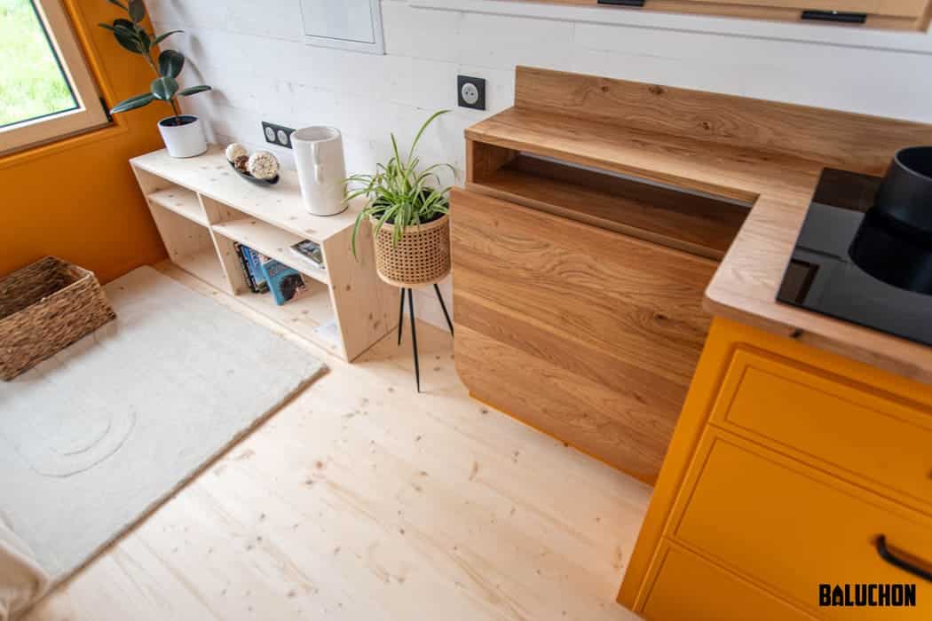 A compact, modern tiny house kitchen with yellow cabinets, wooden countertops, a white shelf with books and decor, a potted plant on a stand, and a woven basket on a light wood floor; sunlight streams in through a window.