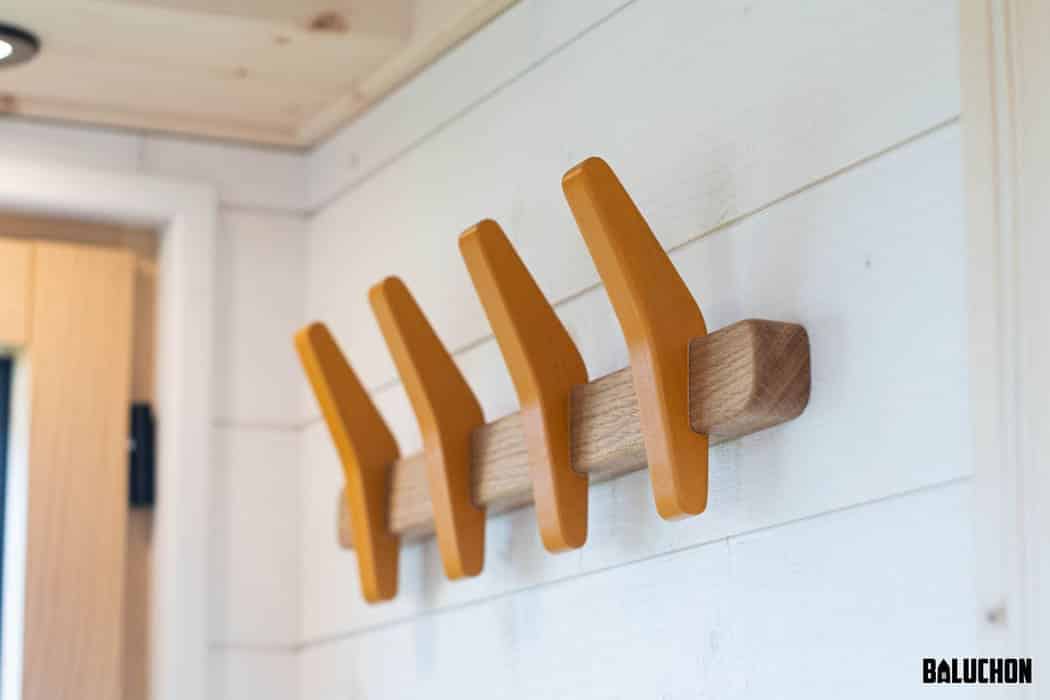 Wall-mounted wooden coat rack with four orange hooks attached to a light-colored plank, perfect for adding cheerful storage to a tiny house, set against a white shiplap wall in a bright room.
