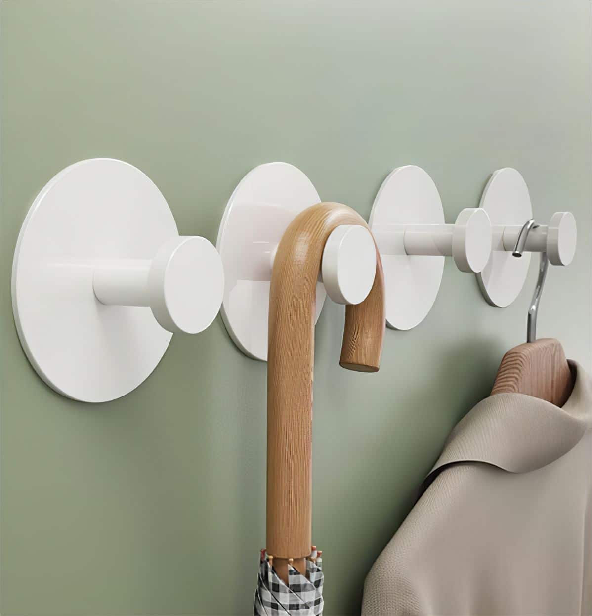 Four round white wall hooks are mounted on a light green wall, creating an organized entryway—ideal for storage renters. An umbrella with a wooden handle and a beige coat hang neatly on two of the hooks.