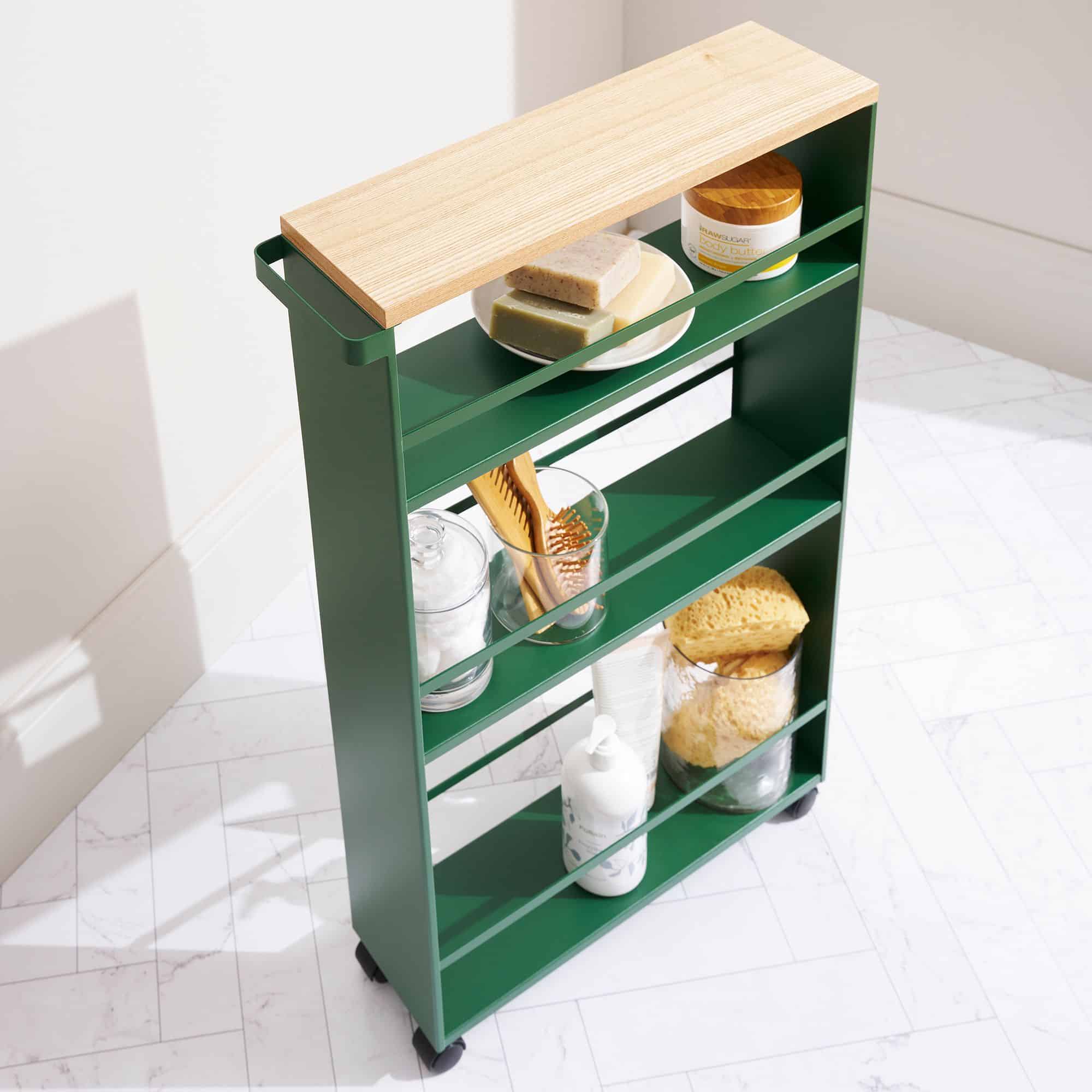 A slim green rolling cart with a wooden top holds jars, soap, a brush, and bottles—an ideal solution for storage renters—placed on a white tiled floor in a well-lit room.