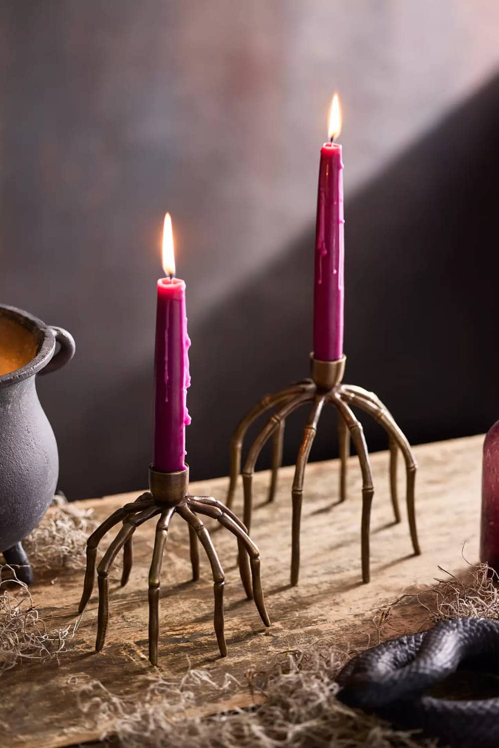 Two lit purple candles stand in holders shaped like bronze skeleton hands on a rustic wooden surface, with a cauldron and decorative moss nearby, making this halloween decor perfect for a spooky atmosphere.