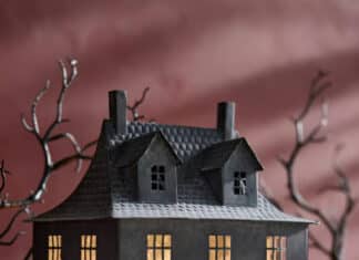 A miniature, dark haunted house with glowing windows sits on a table surrounded by Halloween decor—small pumpkins, dried foliage, and mist—with bare, twisted tree branches in the background.
