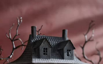 A miniature, dark haunted house with glowing windows sits on a table surrounded by Halloween decor—small pumpkins, dried foliage, and mist—with bare, twisted tree branches in the background.