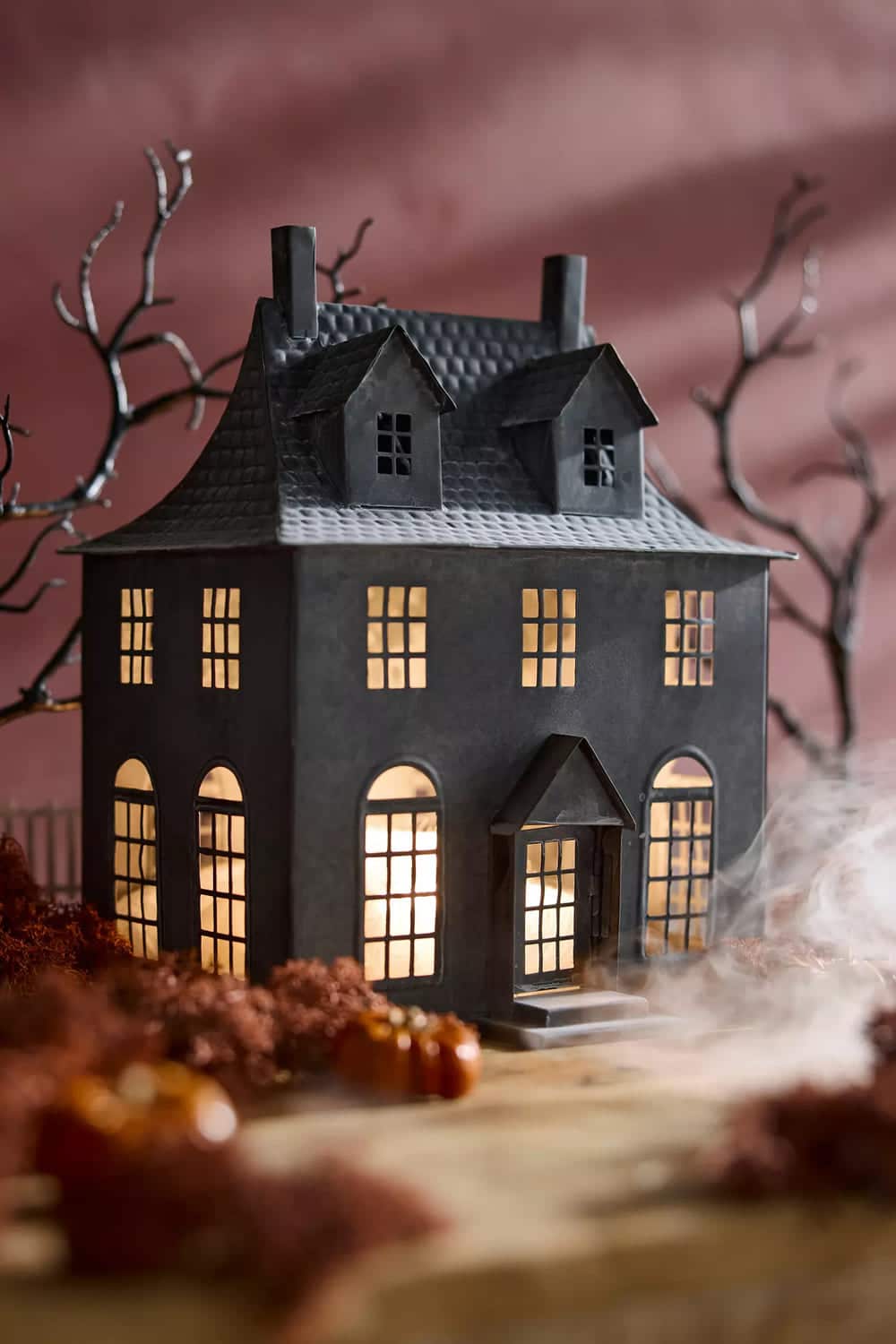 A miniature, dark haunted house with glowing windows sits on a table surrounded by Halloween decor—small pumpkins, dried foliage, and mist—with bare, twisted tree branches in the background.