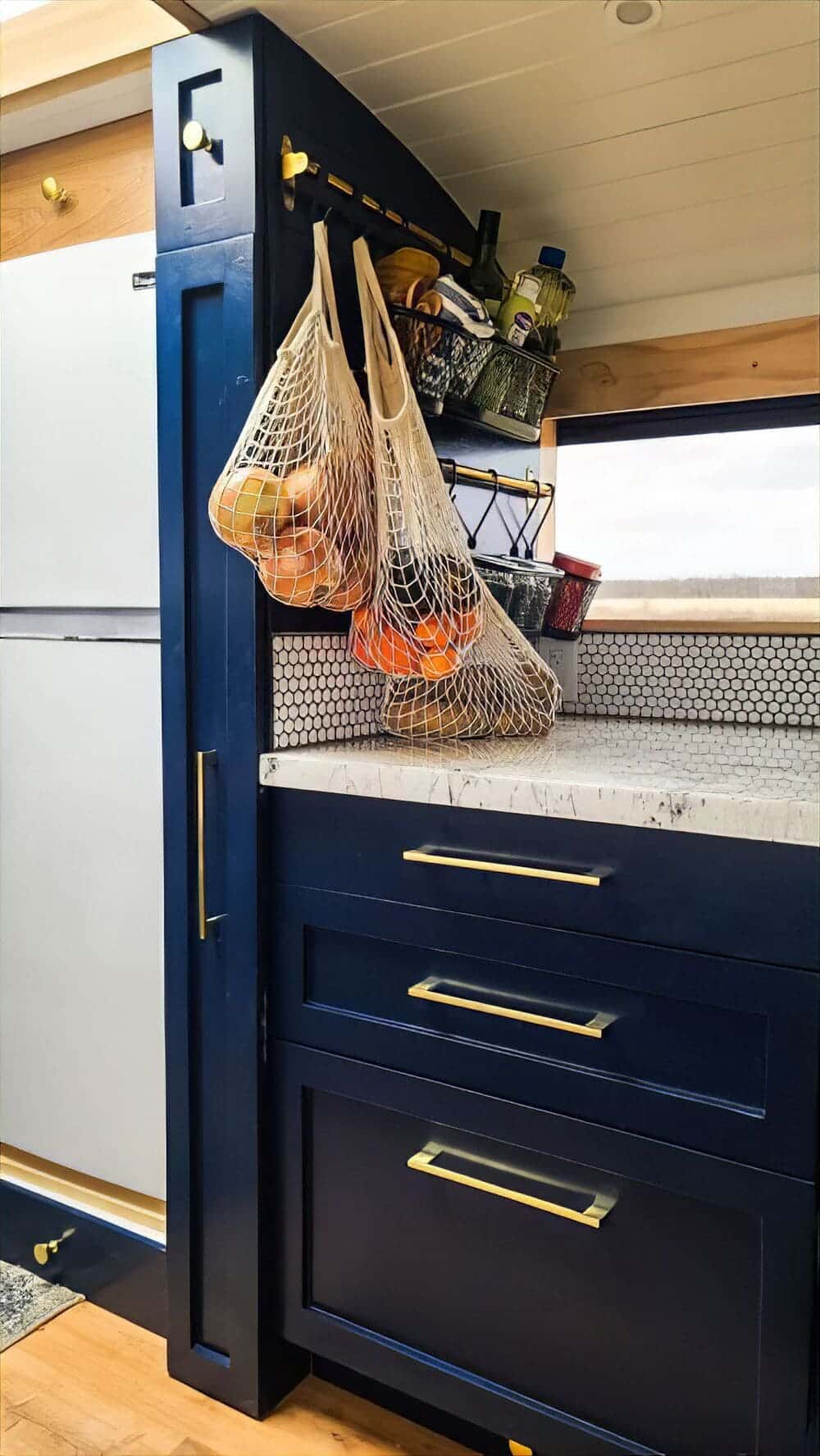 A modern skoolie kitchen with navy cabinets and gold handles, white countertops, small wall shelves with jars and bottles, and mesh bags hanging with onions and oranges. A refrigerator is visible on the left.