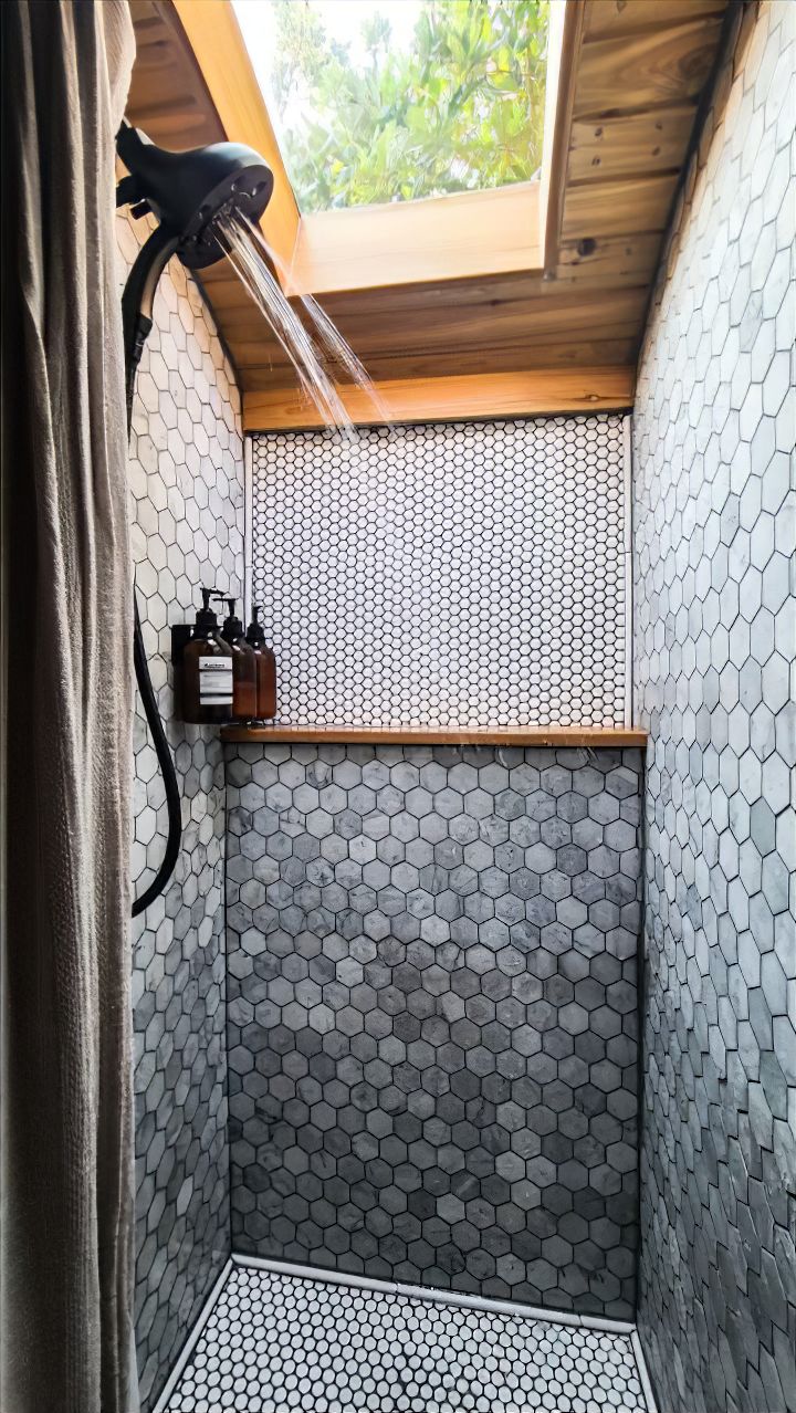 A modern skoolie shower with hexagonal tile walls and floor, a rainfall showerhead, skylight above letting in natural light, and bottles of toiletries neatly arranged on a wooden shelf.