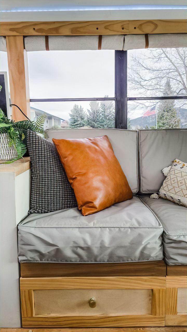 A cozy couch with gray cushions, a brown leather pillow, and two decorative pillows sits next to a window with views of trees and houses, creating the perfect skoolie retreat. A potted plant and drawer are also visible beside the couch.