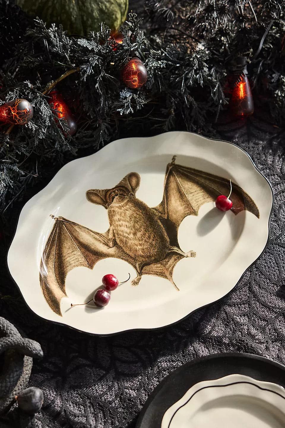 A decorative plate with an illustration of a bat at the center, topped with two cherries. Perfect for Halloween decor, the plate rests on a dark, textured tablecloth surrounded by black foliage and string lights shaped like red berries.