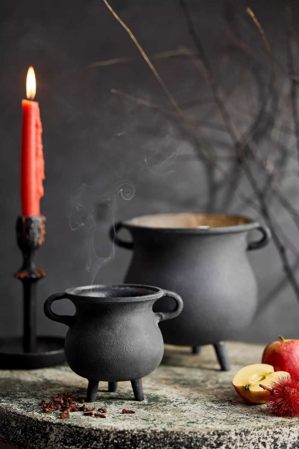 A lit red candle, halloween decor like two black cauldrons—one steaming—and apples are arranged on a stone surface against a dark, moody background with bare branches.
