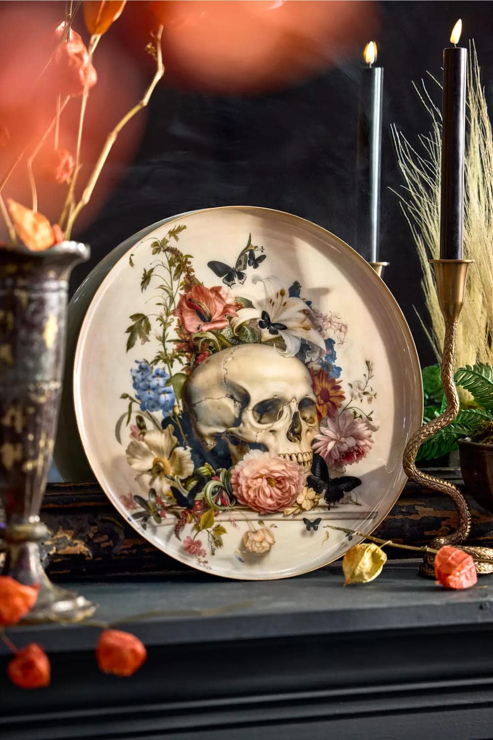 A decorative plate featuring an artistic skull surrounded by colorful flowers and butterflies is displayed on a dark mantel beside black candles and autumnal halloween decor.