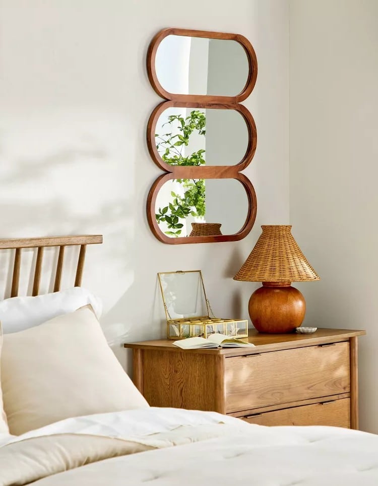 A cozy bedroom features a wooden bed and nightstand, a rattan lamp, an open book, and a glass jewelry box. Three oval mirrors from the Hearth & Hand with Magnolia Fall Collection at Target reflect greenery from a nearby plant.