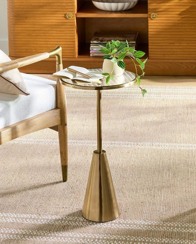 A modern side table from the Target fall collection with a gold base and round top holds a small white vase, books, and reading glasses next to a wooden armchair on a beige rug in a cozy living room.