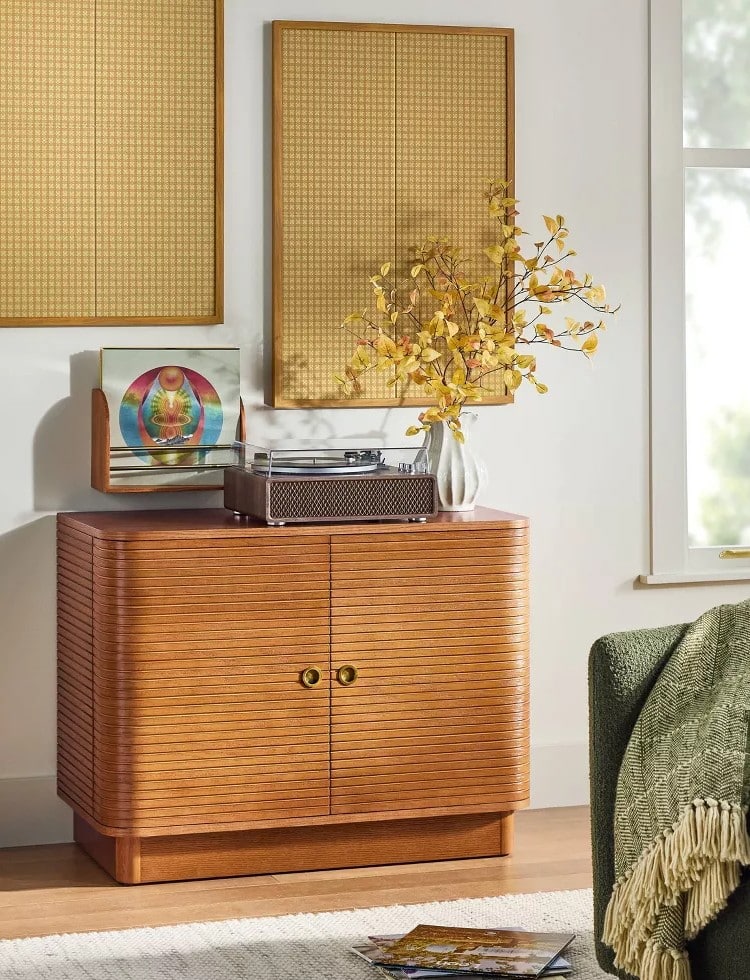 A wooden cabinet with rounded edges holds a turntable, a vinyl record, and a white vase with yellow autumn branches—perfect cozy finds from the Target Fall Hearth & Hand Magnolia Collection. Above, textured gold wall art hangs beside a window and green armchair.