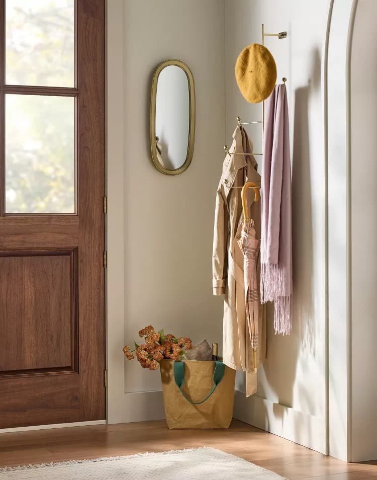A cozy entryway with a wooden door, small oval mirror, yellow beret, coat, pink scarf, and an umbrella hanging on Hearth & Hand hooks. A tan tote bag with flowers sits below as sunlight streams in—a charming Magnolia Collection touch from Target.