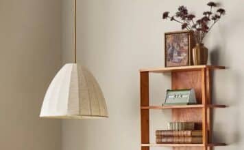 A cozy desk setup with a wooden desk, a fabric pendant lamp from the Hearth & Hand Magnolia Collection at Target, a vintage radio, and books. A wooden wall shelf displays more books, a clock, photo frame, and dried flowers in a vase.