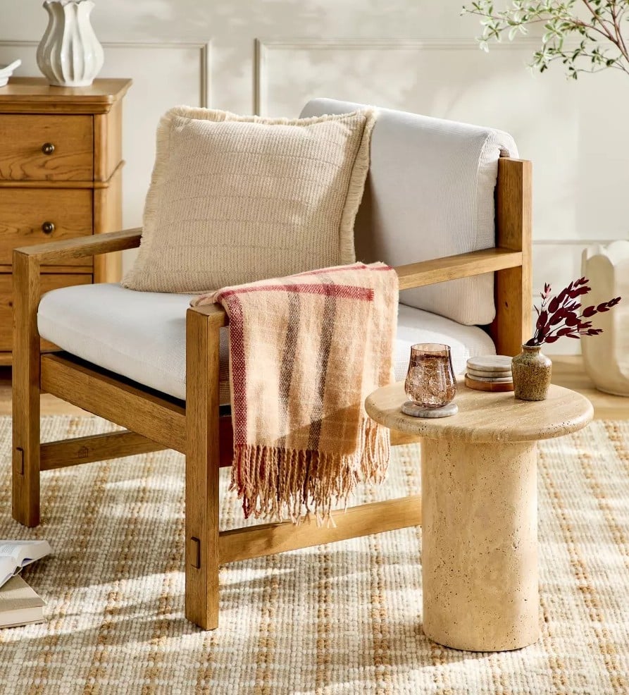 A cozy wooden armchair with white cushions, a beige pillow, and a plaid throw from the Magnolia Collection sits next to a round stone side table with candles and decor in a sunlit, neutral-toned living room.