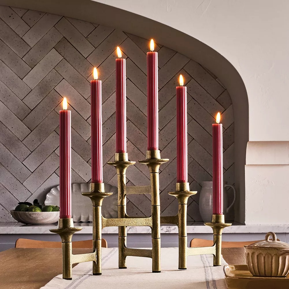 A gold candelabra with six lit red taper candles from the Fall Hearth & Hand Magnolia Collection sits on a dining table. The background features a gray herringbone-patterned backsplash and a bowl of green apples on the left, perfect for Target-inspired décor.