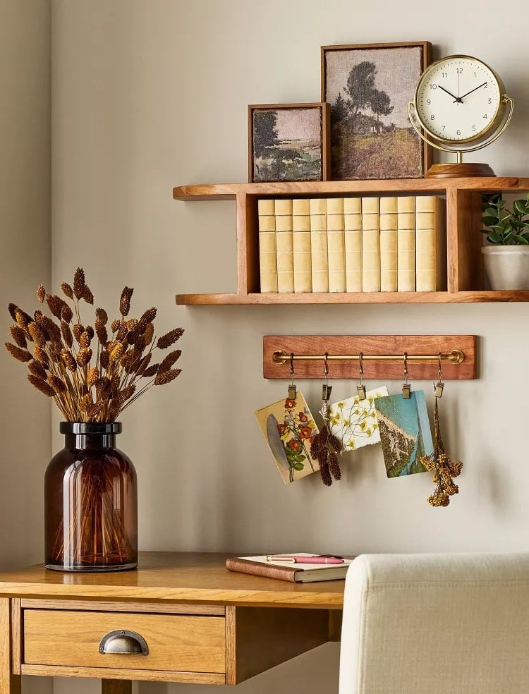 A wooden desk with a brown vase of dried flowers, a notebook, and a beige chair. Above, shelves hold books, framed art, a clock, and Hearth & Hand with Magnolia decor from Target’s Fall Collection displayed alongside postcards and dried flower decorations.