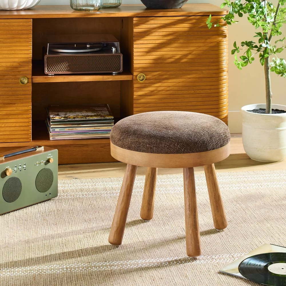 A round wooden stool with a brown cushioned seat from the Hearth & Hand collection sits on a beige rug in front of a wooden cabinet. A potted plant, vintage record player, vinyl records, and a green radio complete the cozy scene.
