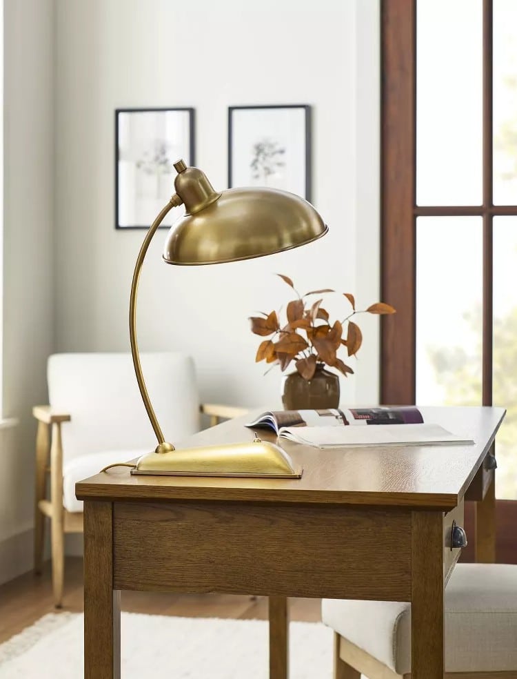 A gold desk lamp sits on a wooden table with an open book and a vase of dried leaves. Cozy home decor shines through, echoing the warm style of the Target fall collection, with light armchair, framed art, and tall windows in the background.
