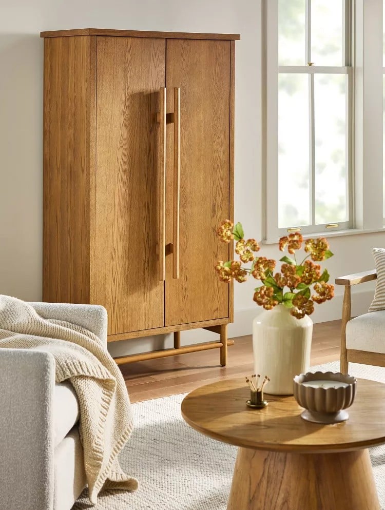 A modern living room featuring a tall wooden cabinet, a round wooden coffee table from the Magnolia Collection at Target, a light gray sofa with a blanket, and large windows letting in natural light.