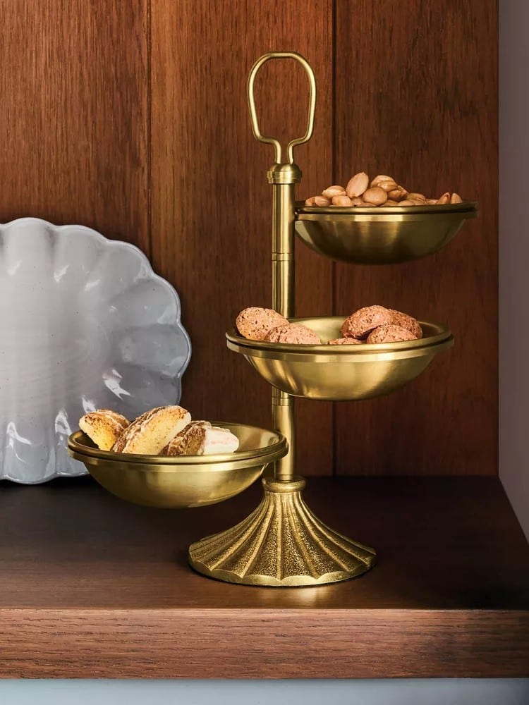 A three-tiered gold serving stand from the Magnolia Collection by Hearth & Hand at Target holds cookies, biscotti, and nuts, displayed on a wooden shelf next to a decorative gray plate.