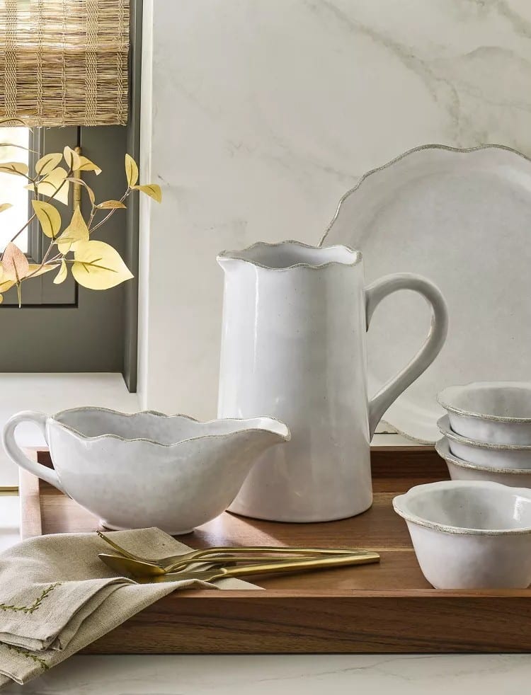 A white ceramic pitcher, gravy boat, bowls, and plates from the Hearth & Hand Magnolia Collection sit on a wooden tray beside folded napkins and gold flatware. Sunlight filters through a nearby window with woven blinds.