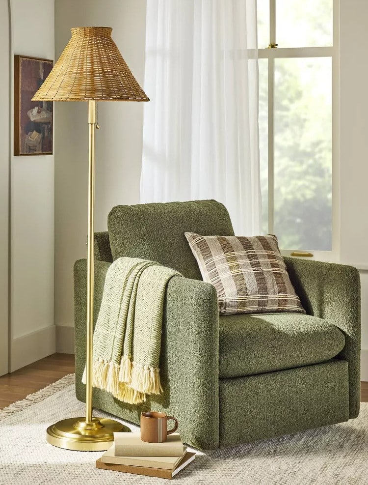 A cozy reading nook with a green armchair, plaid pillow, and throw blanket from the Hearth & Hand with Magnolia Target fall collection. A mug and stacked books sit by a brass floor lamp with wicker shade beside a large window—perfect for cozy chic finds.