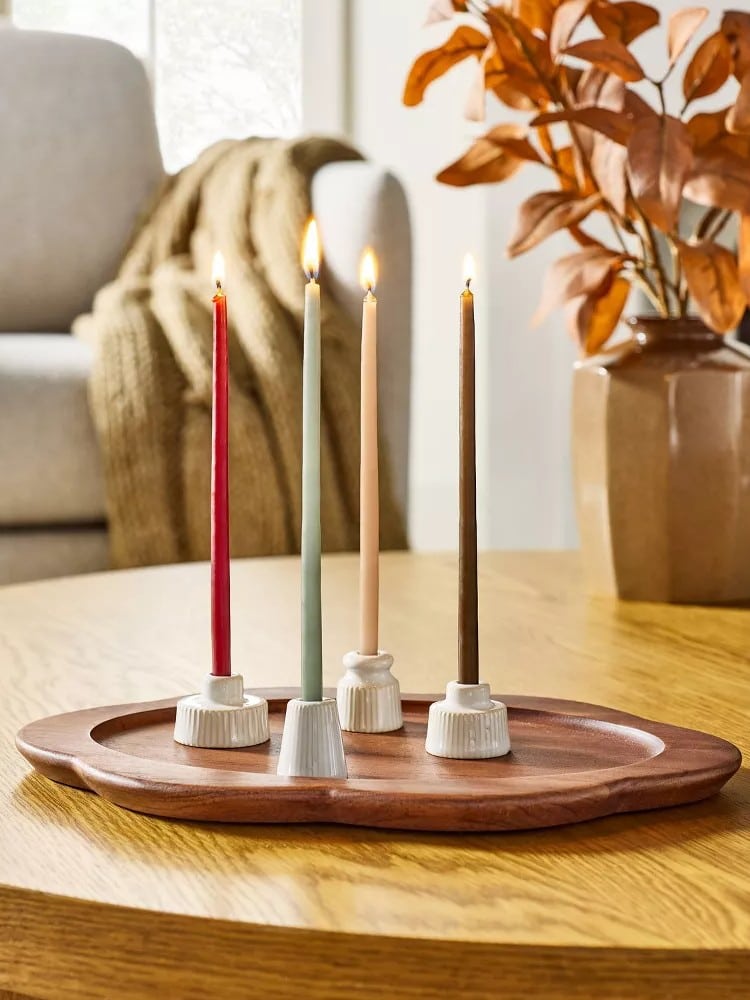 Four lit taper candles in red, green, cream, and brown from the Magnolia Collection stand in white holders on a wooden tray; a vase with orange leaves and a beige sofa add to these cozy chic finds inspired by Target Fall Hearth & Hand.
