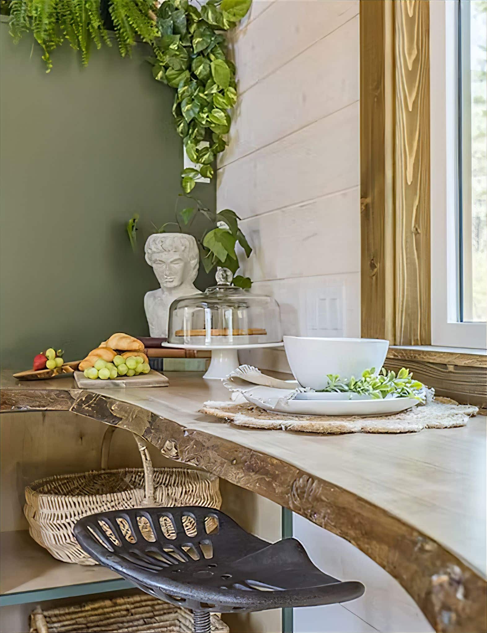 A rustic kitchen corner in a tiny house, featuring a live edge wooden counter, metal stool, plates on wooden chargers, a bowl, glass cake stand, fresh fruits, a classical bust, and green plants hanging near a window.